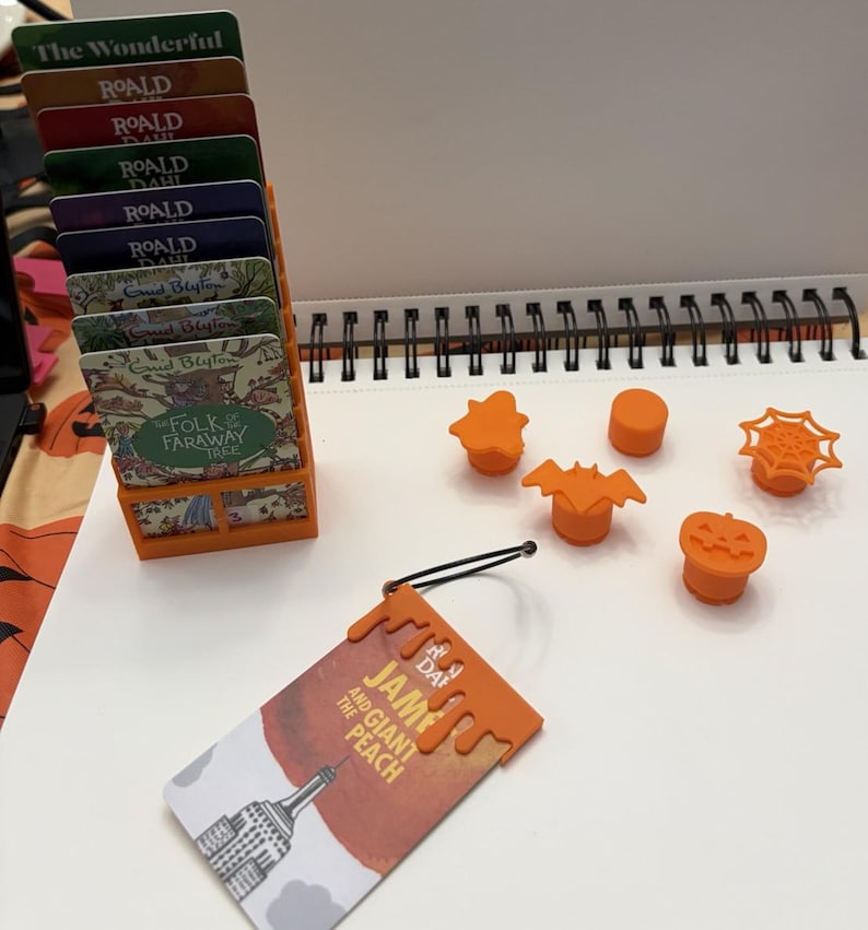 May include: An orange plastic card holder containing several book-themed cards, five small orange plastic toppers in shapes including a bat, pumpkin, and spiderweb, and one orange plastic book tag attached to a card titled James and the Giant Peach.