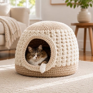 May include: A dome-shaped pet bed made of cream and tan crocheted yarn. The structure features a textured, waffle-like stitch pattern on the upper portion and a smooth, solid base. A brown and white tabby cat rests inside the circular opening.