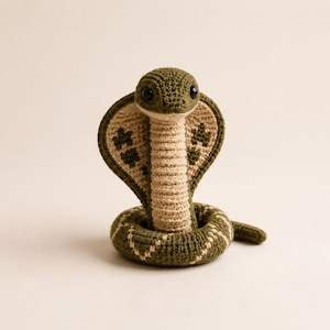 May include: A crocheted cobra toy featuring a coiled body in olive green and cream yarn. The hood displays dark green embroidered patterns, while the front of the neck and belly are cream-coloured. It has two small, round, black plastic eyes.