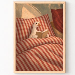 Könnte beinhalten: Ein gerahmter Kunstdruck, der eine weiße Gans mit orangefarbenem Schnabel zeigt, die in einem Bett mit rot-weiß gestreifter Bettwäsche sitzt. Die Gans hält ein aufgeschlagenes Buch mit hellbraunen Seiten. Ein dunkelrotes Kissen ist oben rechts zu sehen.