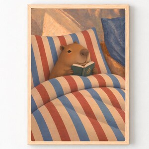 Könnte beinhalten: Ein gerahmter Kunstdruck, der ein braunes Wasserschwein zeigt, das in einem Bett unter einer Decke mit vertikalen roten, weißen und blauen Streifen liegt. Das Wasserschwein hält ein kleines, offenes grünes Buch mit einem goldenen Blattsymbol.