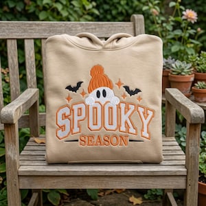 May include: A beige hooded sweatshirt featuring an embroidered design of a white ghost wearing an orange beanie, surrounded by black bats and orange stars, with the text SPOOKY SEASON embroidered in orange and white block lettering.