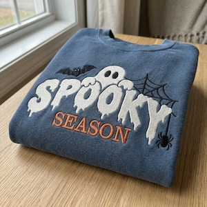 May include: A blue crewneck sweatshirt featuring white embroidered text that reads SPOOKY with a ghost, a black bat, a spider web, and a small spider. The word SEASON is embroidered in orange below the ghost. The design is centered on the chest.