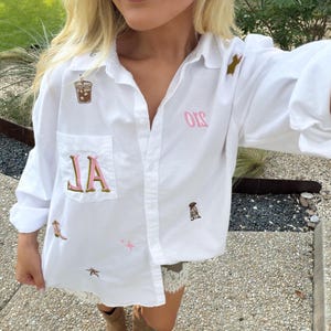 May include: A white button-down shirt featuring various embroidered patches including a coffee cup, a pink lettered monogram on the chest pocket, a cowboy boot, a star, and other small decorative icons scattered across the fabric surface.