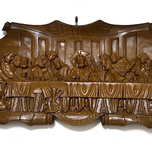 May include: A brown wooden wall plaque featuring a relief carving of The Last Supper. The scene depicts thirteen figures seated at a long table with a draped cloth. The carving is framed by a scroll-like border with curled edges at the sides.