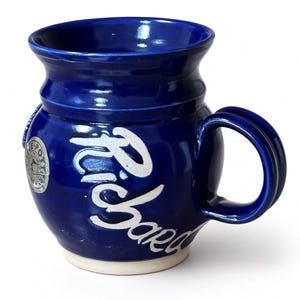May include: A glossy cobalt blue ceramic mug with a rounded body and a flared rim. The name Richard is written in white script across the front. A circular silver-toned emblem with the text LE POTTERY is visible on the side near the handle.