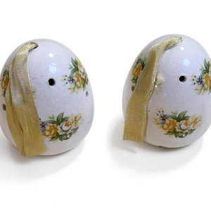May include: Two white ceramic egg-shaped ornaments featuring a delicate floral pattern with yellow roses and green leaves. Each egg has a small circular hole near the top and a gold-coloured ribbon loop attached for hanging purposes.