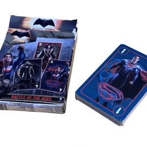 May include: A deck of Batman v Superman: Dawn of Justice playing cards featuring a box with character illustrations and the text BATTLE OF THE AGES. The cards display Superman with a red and blue logo and the number one on a dark blue background.