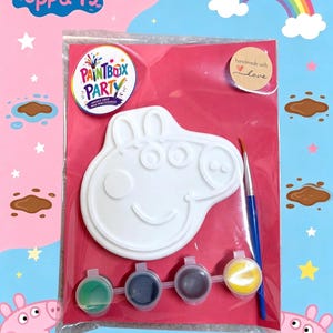Pode incluir: Um kit de pintura com o tema Peppa Pig numa embalagem de plástico transparente sobre um cartão rosa. O kit inclui uma figura de gesso branca em forma de Peppa Pig, um pincel azul e quatro pequenos potes de tinta nas cores verde, azul, castanho e amarelo.