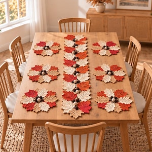 May include: A rectangular table runner and six circular placemats made of crocheted leaves in shades of red, orange, and cream. The runner and placemats are decorated with small, dark brown pom-poms arranged in clusters throughout the leaf patterns.