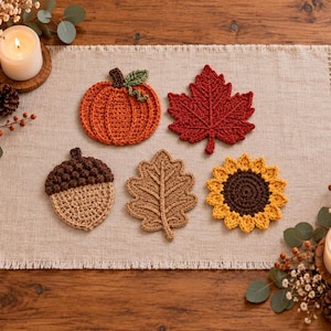 Könnte beinhalten: Ein Set aus fünf gehäkelten Untersetzern mit herbstlichen Motiven, darunter ein orangefarbener Kürbis mit grünem Stiel, ein dunkelrotes Ahornblatt, eine braune Eichel, ein beiges Eichenblatt und eine gelbe Sonnenblume mit dunkelbrauner Mitte.