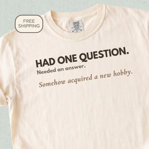 May include: A cream-colored short-sleeve t-shirt featuring dark brown text that reads: HAD ONE QUESTION. Needed an answer. Somehow acquired a new hobby. The shirt has a crew neckline and a Comfort Colors brand label visible at the inner collar.