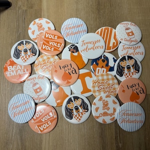 May include: A collection of round pinback buttons featuring orange and white designs. Graphics include a dog wearing heart-shaped glasses, text reading 'Vols', 'Tennessee Volunteers', 'Home Sweet Home', 'Lucky to be a Vol', and 'Beat Em All'.