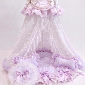 May include: A lavender pet bed featuring a canopy with white lace, ruffles, and pearl strands. The bed includes a round ruffled pillow, a bone-shaped pillow with the name "chloe" embroidered in white, and several satin bows throughout the design.