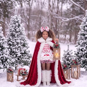 May include: A deep red velvet cape with white faux fur trim, worn over a white and red candy cane striped dress with ruffled layers. The outfit includes a matching oversized hair bow and white faux fur leg warmers, paired with white boots.