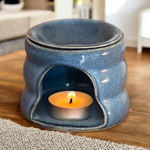May include: A blue ceramic oil burner with a speckled finish and a rounded, wavy silhouette. The piece features a wide top basin for wax or oil and a large arched opening at the base containing a small, lit tea light candle with a warm glow.