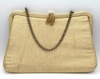 May include: A gold-toned, shimmering fabric clutch bag with a structured frame and a thin, metallic chain shoulder strap. The bag features a decorative, leaf-shaped metal clasp at the centre top, providing a secure closure for the main compartment.