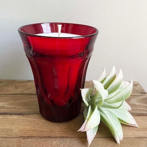 May include: A red glass candle holder with a white candle inside. The candle holder has a textured design and is sitting on a wooden surface.