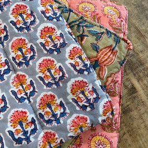 May include: Three fabric placemats with a floral pattern. The top placemat is gray with blue and pink flowers. The middle placemat is green with pink and yellow flowers. The bottom placemat is pink with yellow and red flowers.