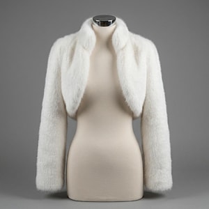 Puede incluir: Un bolero de piel sintética blanca con mangas largas y cuello alto. La prenda corta se exhibe sobre un maniquí, mostrando un acabado de pelo suave y texturizado que cubre toda la superficie exterior de la pieza, desde el hombro hasta el puño.
