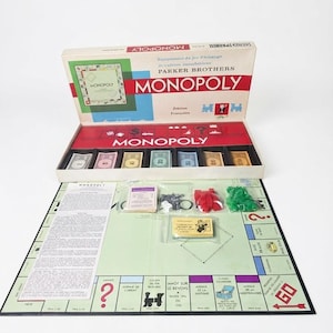 May include: A Monopoly board game set featuring an open box with paper money in compartments, a game board with French text, and plastic game pieces including houses, hotels, and tokens in sealed plastic bags, all displayed on a white surface.