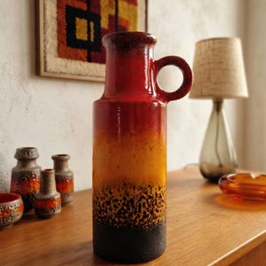 Puede incluir: Un jarrón de cerámica alto con cuerpo cilíndrico y una sola asa lateral circular. El esmalte presenta un degradado que va desde una base de color marrón oscuro con textura hasta tonos vibrantes de amarillo, naranja y rojo en el borde.