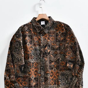 May include: A long-sleeve button-down shirt featuring a complex patchwork pattern in shades of black, dark orange, and cream. The garment includes a standard collar, a single chest pocket, and a small rectangular black label with white text.