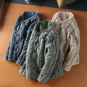 May include: Three quilted jackets in dark blue, sage green, and beige. Each jacket features a collarless round neckline, a button-front closure, and long sleeves. The fabric has a soft, textured, wavy quilted pattern throughout the exterior.