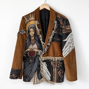 May include: A brown corduroy blazer featuring a tapestry-style panel on the left side depicting a figure with a halo holding a red heart. The garment includes frayed edges, black lining, and a right-side panel with white wing imagery and ornate patterns.