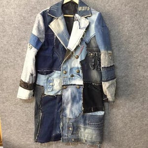Op de afbeelding: Een lange jas opgebouwd uit diverse blauwe denim stofstukken. Het kledingstuk is voorzien van een reverskraag, lange mouwen en een dubbele rij metalen knopen aan de voorzijde. Het ontwerp heeft duidelijk zichtbare gerafelde randen.