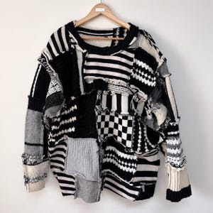 May include: A long-sleeve sweater constructed from various black, white, and gray knit fabric patches. The garment features a mix of patterns including horizontal stripes, a checkerboard design, and textured cable knit sections with raw, frayed edges.