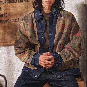 Könnte beinhalten: Ein Lagen-Outfit bestehend aus einer dunkelblauen Jeansjacke mit Metallknöpfen über einem braunen Hemd, kombiniert mit einer gemusterten Außenjacke mit geometrischen Designs in Braun-, Beige- und Rottönen mit langen Ärmeln und Kragen.
