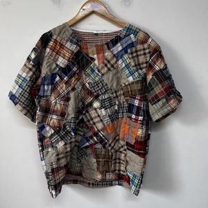May include: A short-sleeved shirt constructed from a patchwork of various plaid and checkered fabric squares in shades of brown, blue, red, white, and yellow. The garment features a round neckline and is displayed on a wooden clothes hanger.