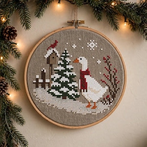 Peut inclure: Un tambour à broder circulaire contenant un motif au point de croix sur tissu beige. La scène représente une oie blanche avec une écharpe rouge, un sapin enneigé, un nichoir avec un oiseau rouge et des branches de baies rouges.