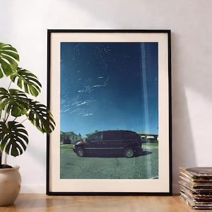 Può includere: Una stampa artistica da parete verticale incorniciata che raffigura un minivan di colore scuro parcheggiato in una strada residenziale sotto un cielo blu intenso. L'immagine ha un'estetica vintage con striature di luce e texture.