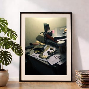 May include: A framed art print featuring a cluttered desk scene with a gold chain, a small pipe, a prescription bottle, and papers labeled Lamar and Section 80. The composition includes books and various small objects arranged in a dimly lit setting.