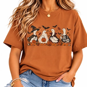 May include: A rust-colored short-sleeve t-shirt featuring a graphic print of four ducks dressed in Halloween costumes. The ducks are depicted as a witch, a ghost, a vampire, and a skeleton, surrounded by autumn leaves, pumpkins, and flying bats.
