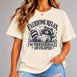 May include: A cream-colored short-sleeve t-shirt featuring a dark blue graphic of a raccoon wearing sunglasses, lying down with hands behind its head. The text reads "EVERYONE RELAX I'M PROFESSIONALLY DEVELOPED" with small star and flower accents.