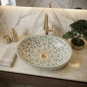 Puede incluir: Un lavabo ovalado de cerámica con un patrón floral en verde salvia y blanco que presenta un diseño de estrella geométrica central. El lavabo incluye un desagüe dorado y se acompaña de un grifo dorado, un dispensador de jabón y una toalla doblada.