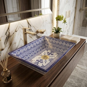 May include: A rectangular ceramic vessel sink featuring a white base with intricate blue geometric and floral patterns. The sink includes a gold-toned drain and is paired with a matching gold-toned faucet, set against a marble wall background.