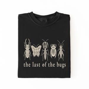 Puede incluir: Una camiseta negra de cuello redondo con un estampado gráfico en color crema de cinco insectos diferentes, incluyendo un escarabajo, una mariposa, una mantis y un saltamontes, con el texto "the last of the bugs" impreso debajo.