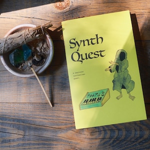 May include: A yellow book titled Synth Quest with a subtitle A beginner synthesizer quest. The cover features a green illustration of a hooded figure and a synthesizer. Next to the book is a small bowl containing a bundle of sage and blue stones.