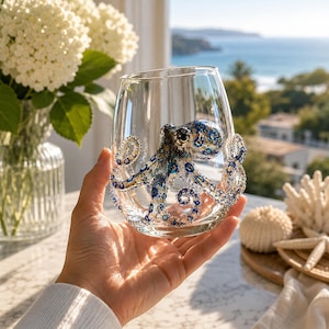 Op de afbeelding: Een helder drinkglas met een driedimensionale octopusdecoratie. De octopus is uitgevoerd in blauwe, zwarte en witte tinten, met ingewikkelde patronen op de kop en acht gekrulde tentakels die zich rondom het oppervlak van het glas bevinden.
