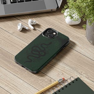 Puede incluir: Una funda de smartphone verde oscuro con la silueta negra de una serpiente enroscada rodeada de sutiles ilustraciones de hojas botánicas. La funda tiene un recorte para una cámara de doble lente y está sobre una superficie de madera clara.