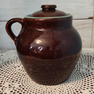 May include: A dark brown ceramic pot with a matching lid and a single side handle. The pot features a smooth, glossy finish and a light green rim accent visible where the lid meets the body. The vessel has a rounded, bulbous shape.