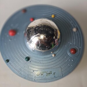 May include: A circular blue solar system model featuring a central chrome dome, various colored spheres representing planets, and concentric orbital rings. A rectangular label at the bottom displays the text "GREEN COUNTY BANK" and "365 DAYS".