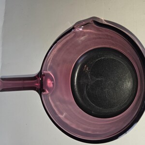 May include: A translucent purple glass saucepan with a circular bowl and a single handle extending from the side. The interior base of the pan is dark and circular, while the rim features a small pouring spout on the side opposite the handle.