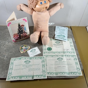 May include: A Cabbage Patch Kids doll with light skin, blue eyes, and curly light brown yarn hair. The doll is accompanied by a greeting card, a small decorative emblem, and several official birth certificate and adoption paper documents.