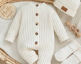Puede incluir: Un pelele de manga larga en punto acanalado color crema con cinco botones marrones en la parte delantera, un gorro a juego con un pequeño parche rectangular marrón, patucos de punto acanalado y un letrero de madera con el texto Hello world.