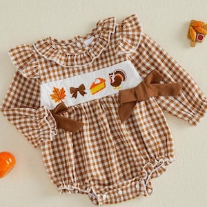 Puede incluir: Un pelele de manga larga con estampado de cuadros vichy en marrón y blanco, con un panel frontal blanco fruncido y bordado con motivos otoñales que incluyen una hoja de arce, un lazo, una porción de tarta de calabaza y un pavo. Hombros con volantes y lazos marrones.